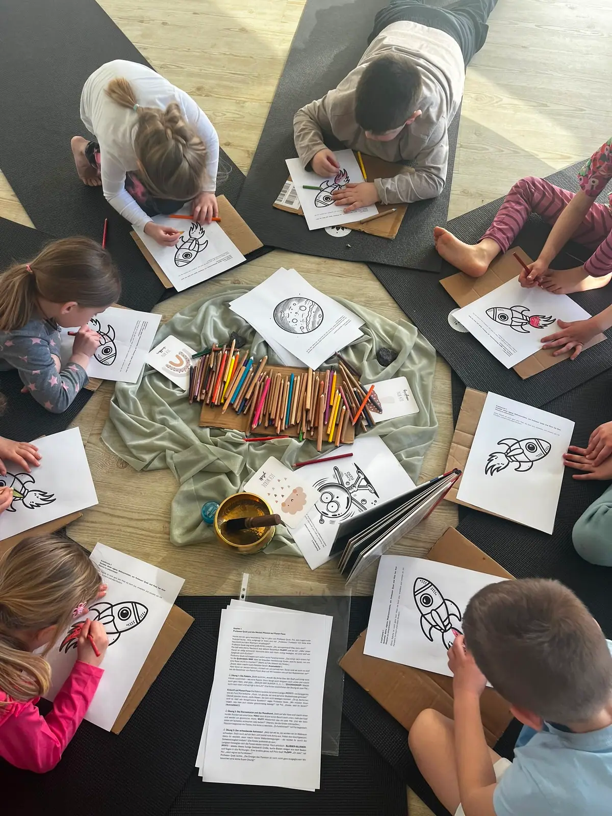 Kinder liegen im Kreis auf Matten und malen Raketenbilder aus, in der Mitte Buntstifte und eine Klangschale