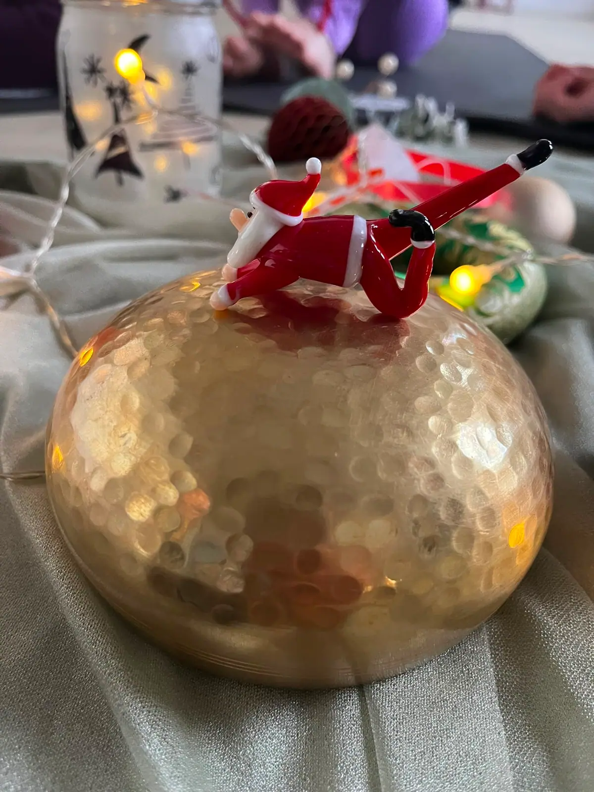 Ein gläserner Weihnachtsmann liegt ausgestreckt auf einer goldenen Klangschale, dahinter eine Lichterkette